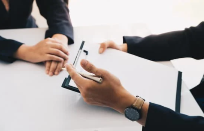 Picture of hands lying on top of a table and another pair of hands holding a clipboard and a pen.